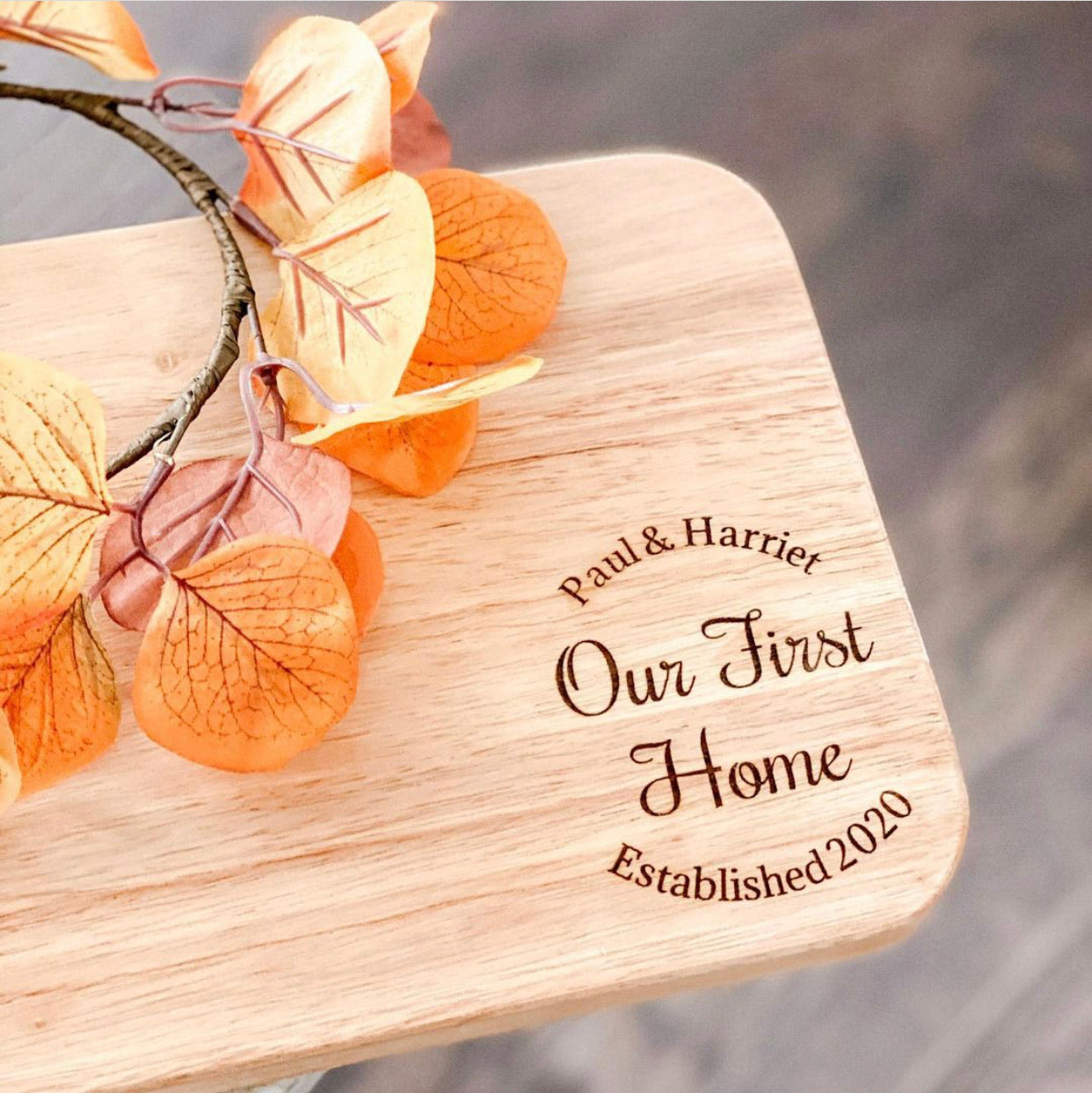 A wooden charcuterie board with engraved text 'Our First Home' and 'Established 2020', displayed with decorative leaves on top.